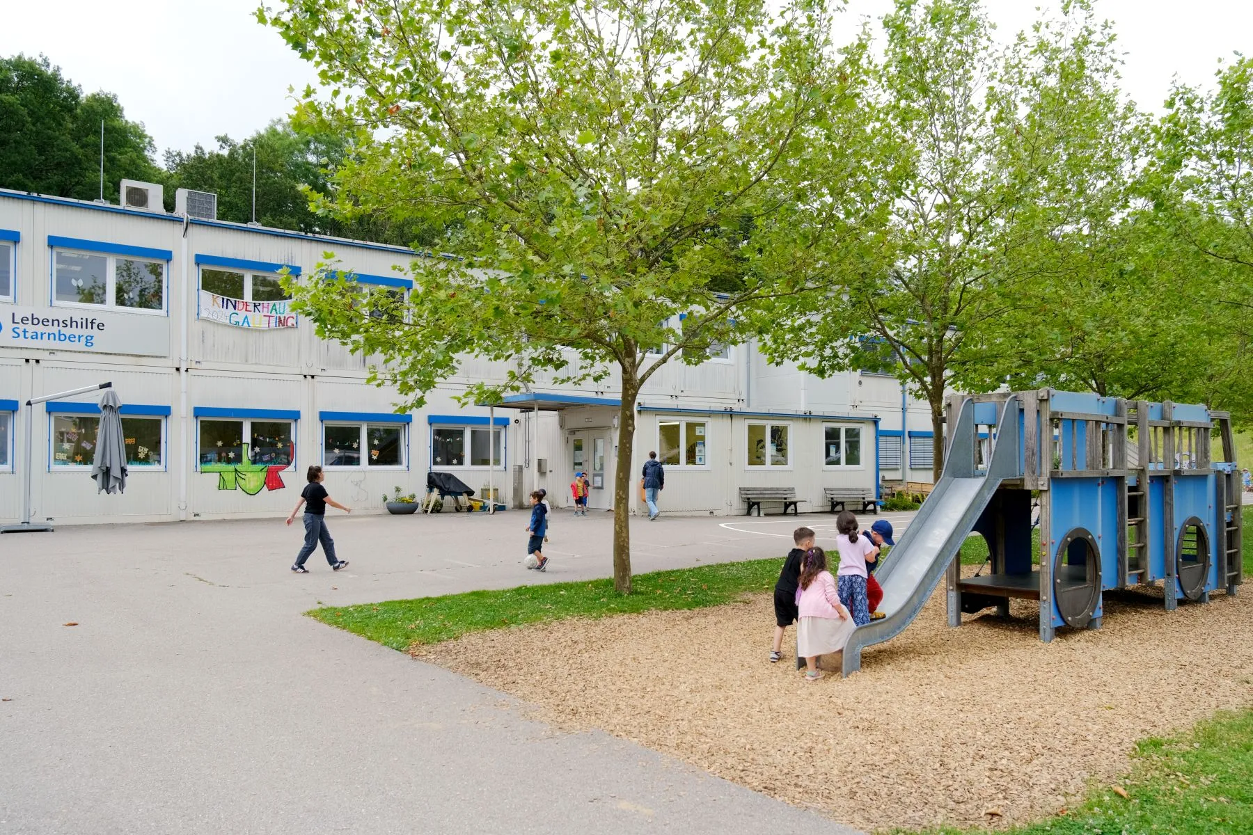 Viele Kinder spielen im Garten, es gibt eine Rutsche, manche spielen Fu&szlig;ball, im Hintergrund sieht man das Kinderhaus Gauting