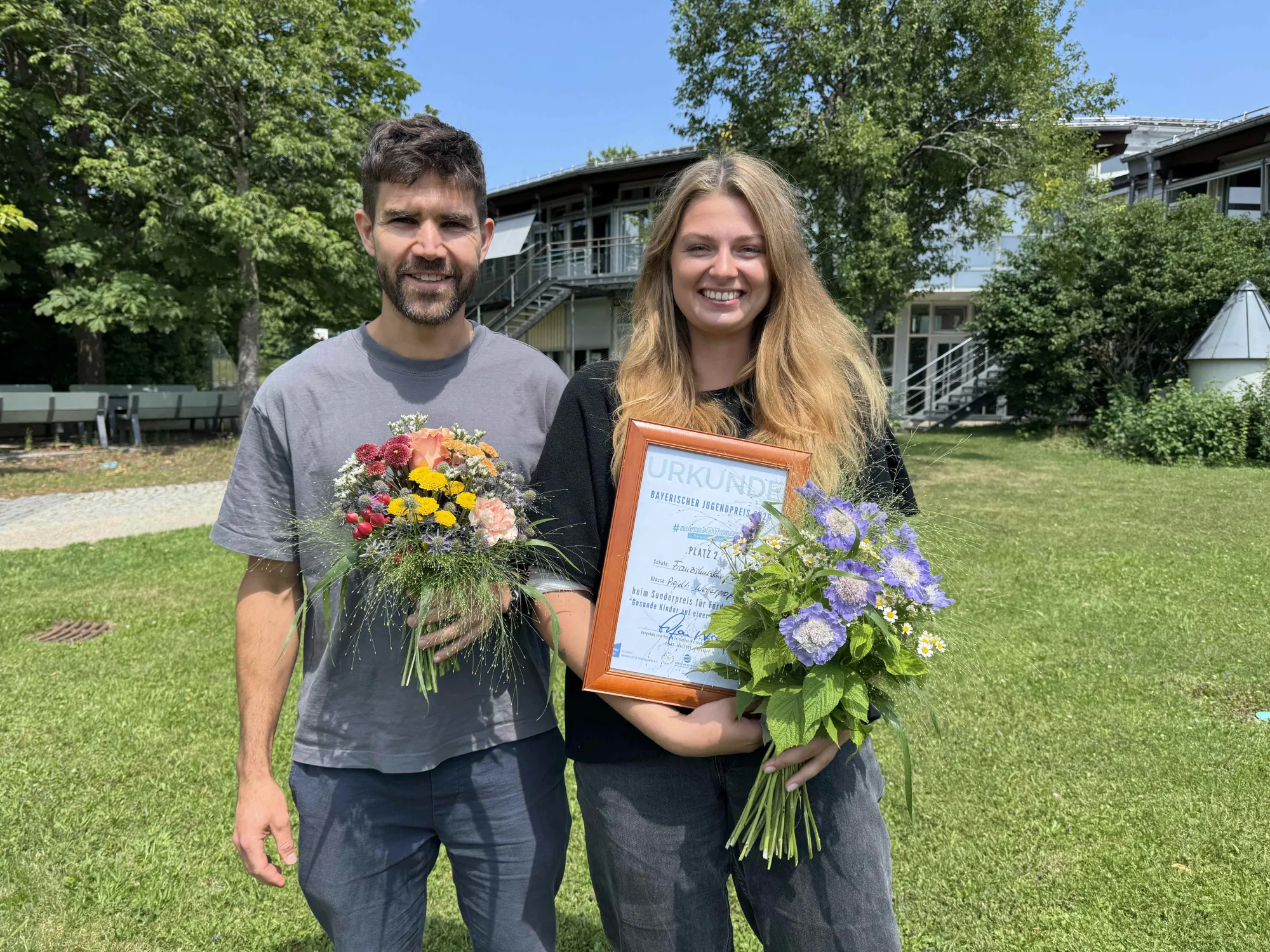 Schulleiter Alexander Sali und Schulsozialpädagogin Carly Neteler der Franziskus-Schule halten je einen Blumenstrauß und gemeinsam die Urkunde über den Bayerischen Jugendpreis 2026 in den Händen.