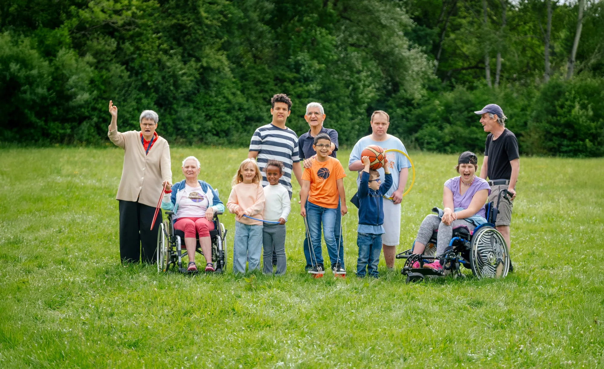 Eine kleine Menschengruppe steht auf einer Wiese, alle lachen und spielen miteinander, 2 Menschen im Rollstuhl